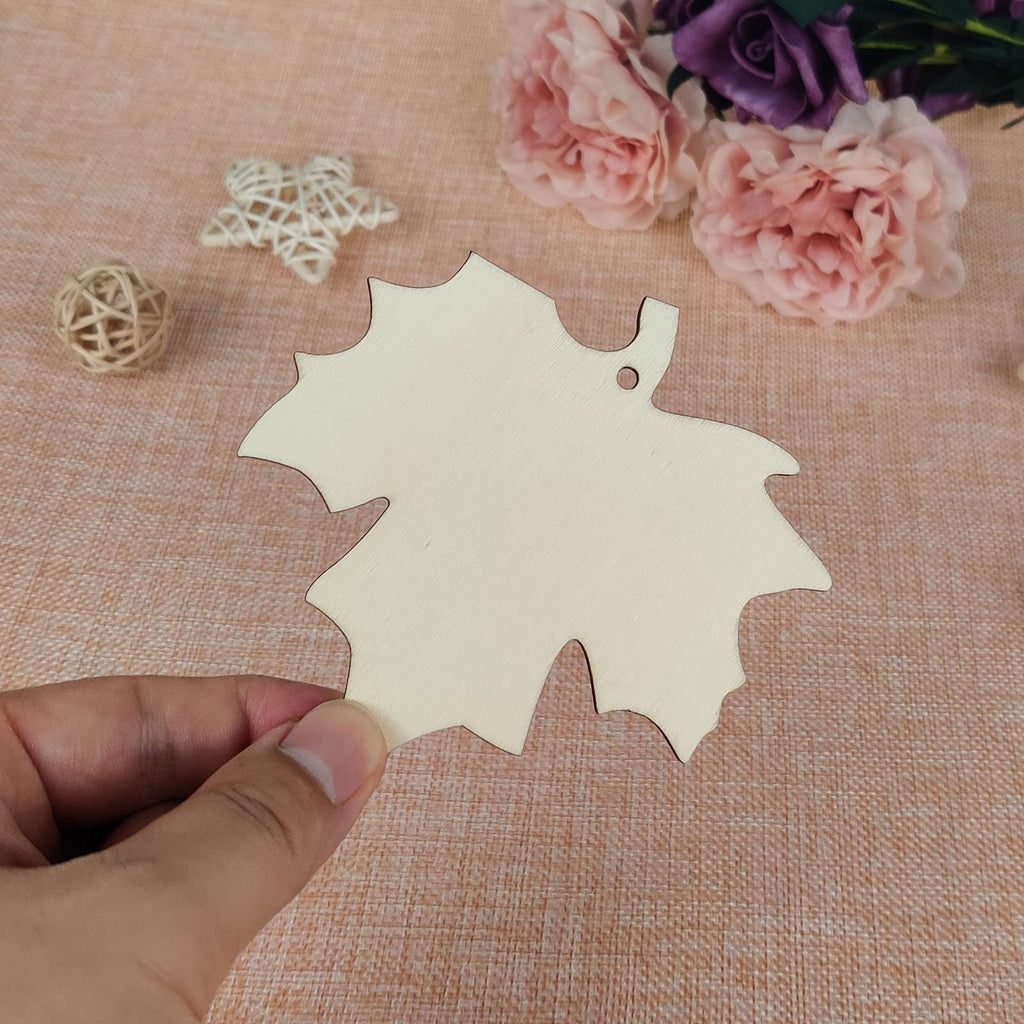 Hand holding a light-colored wooden maple leaf cutout with a hole for hanging. Decorative elements and flowers in the background.