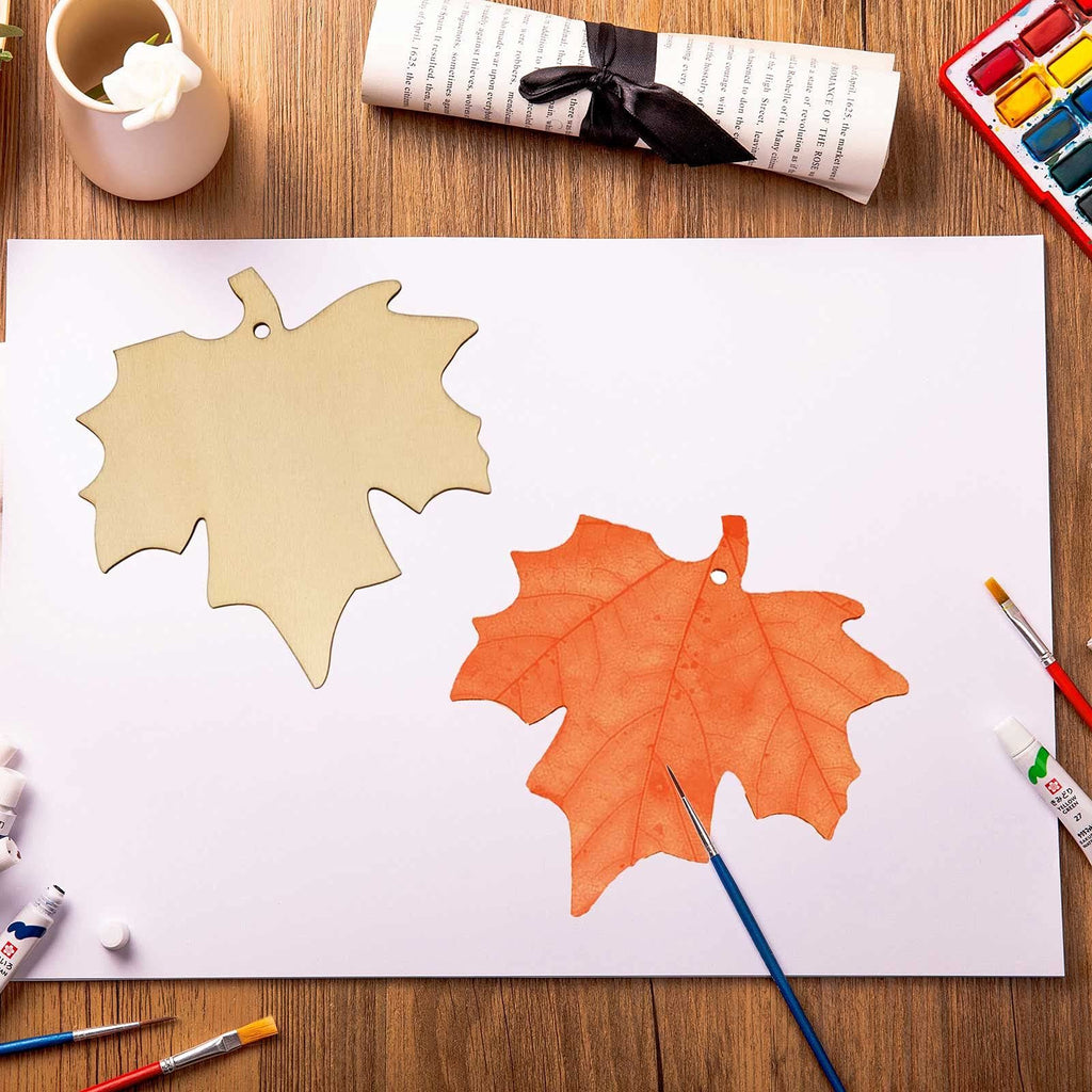 Two maple leaf wood cutouts, one natural wood and one orange, on a white surface with art supplies.