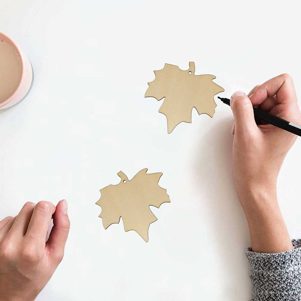 Two maple leaf wood cutouts on a white surface, one being drawn on with a black marker by a hand.