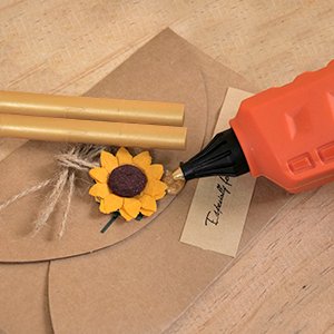 Orange cordless hot glue gun dispensing glue onto a small tag attached to a kraft paper envelope with two gold glue sticks and a felt sunflower decoration.