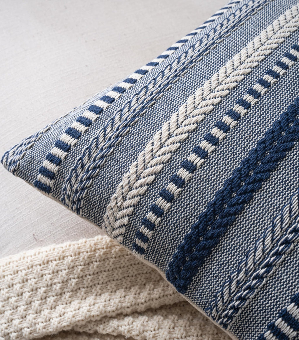 Close-up of a navy blue and white woven throw pillow cover with textured stripes and a cream knit blanket.