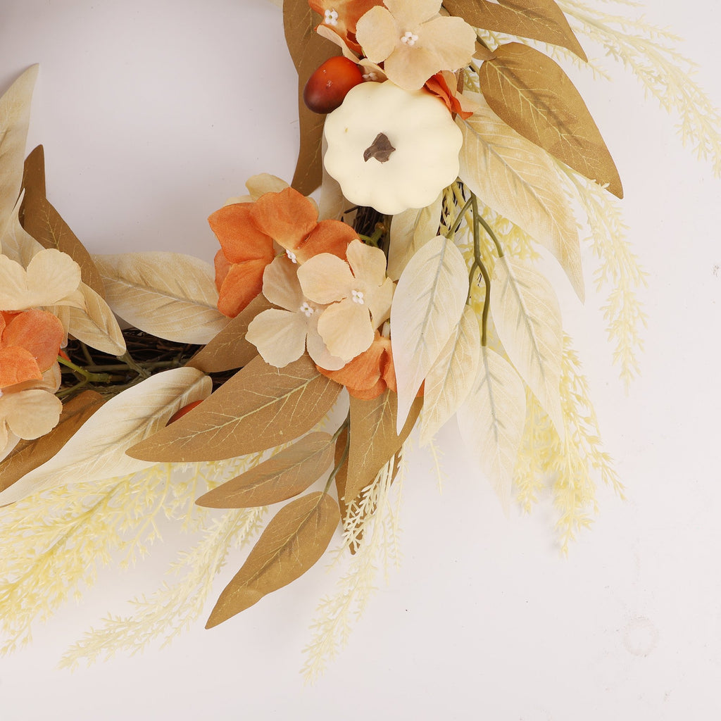 Close-up of an artificial autumn wreath featuring a white pumpkin, orange and beige flowers, and brown and cream leaves against a white background.