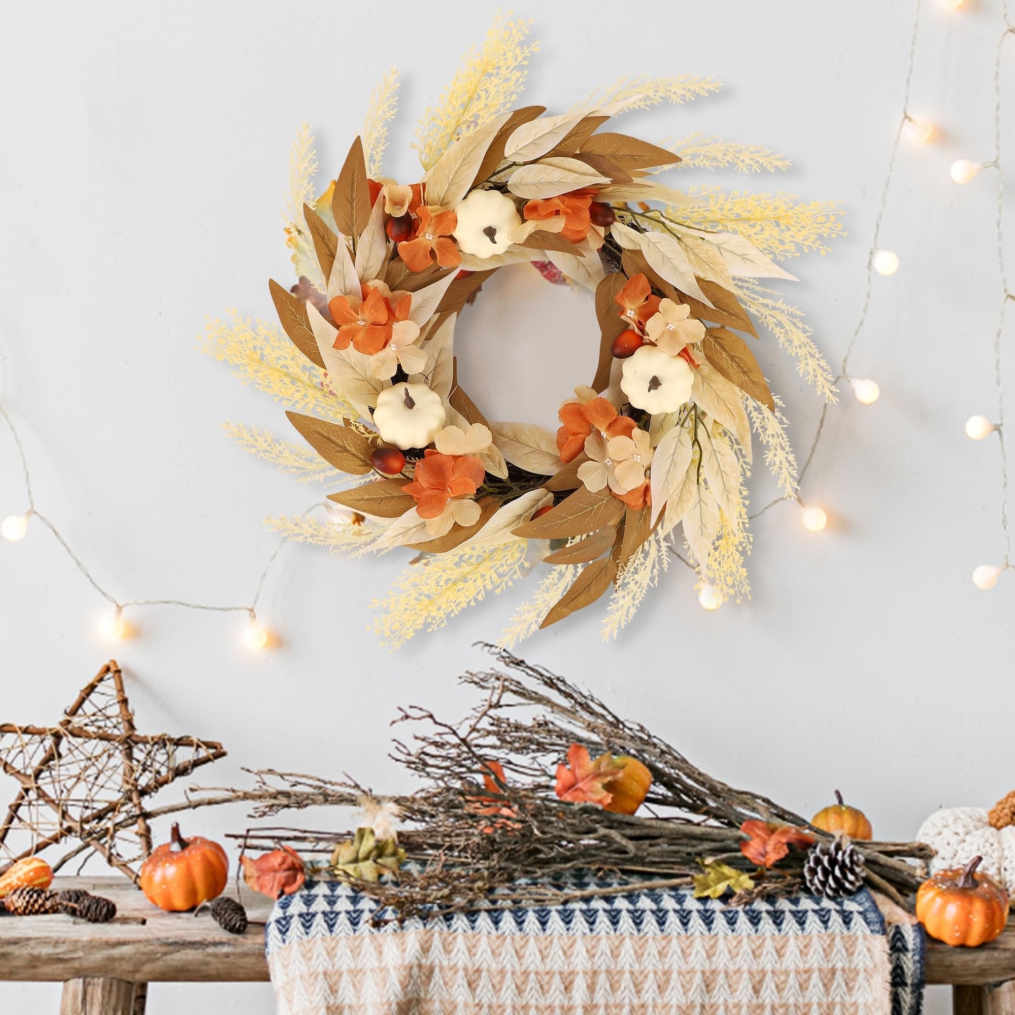 Artificial fall wreath with white pumpkins, orange flowers, and autumn leaves hanging on a wall with string lights.