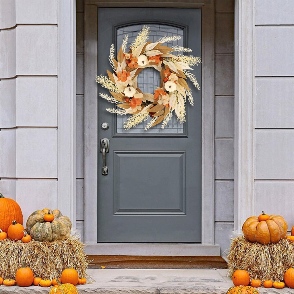 Artificial fall wreath with white pumpkins, orange flowers, and golden leaves hanging on a gray front door, flanked by hay bales with pumpkins.