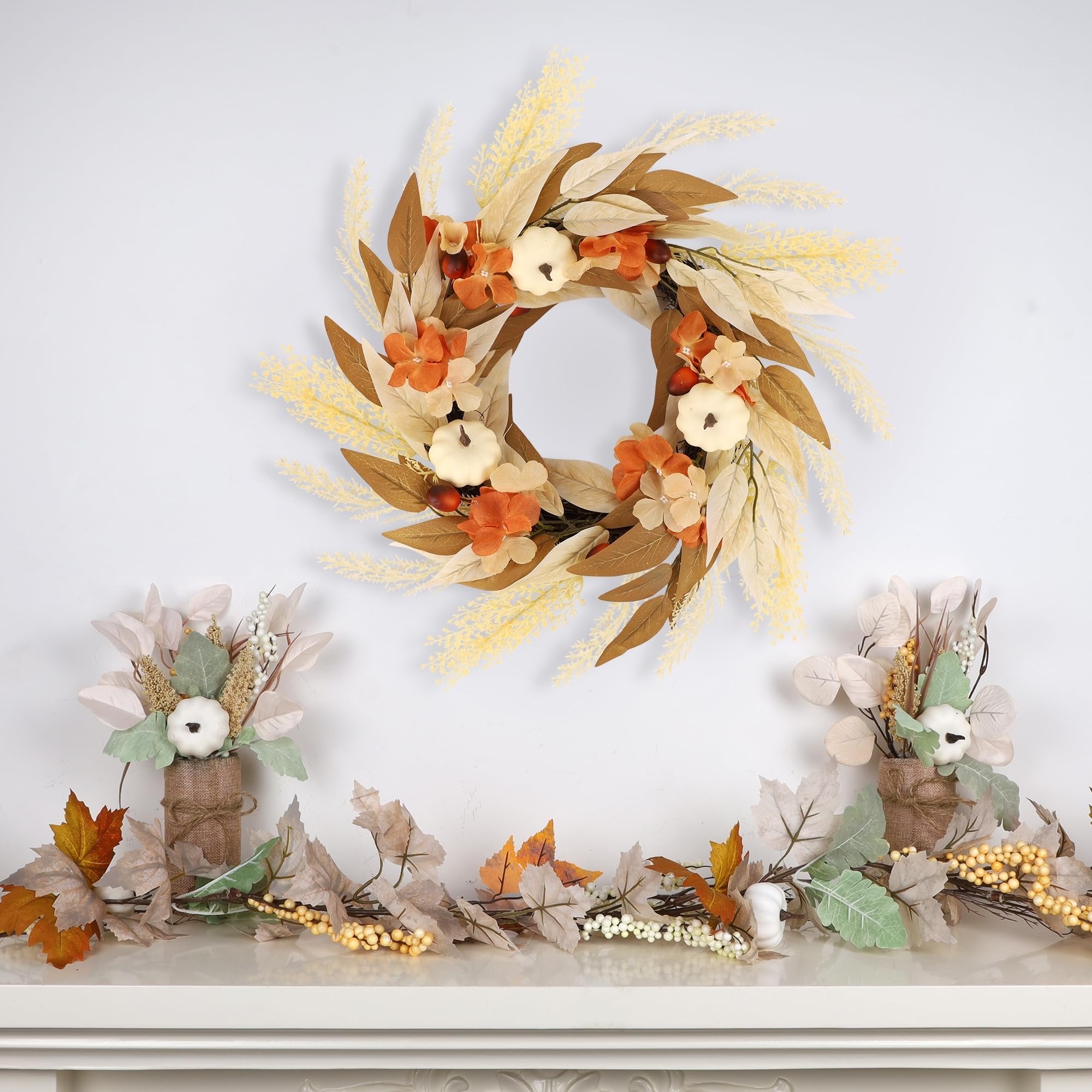 Artificial fall wreath with white pumpkins, orange flowers, and autumn leaves, displayed above a garland on a mantelpiece.
