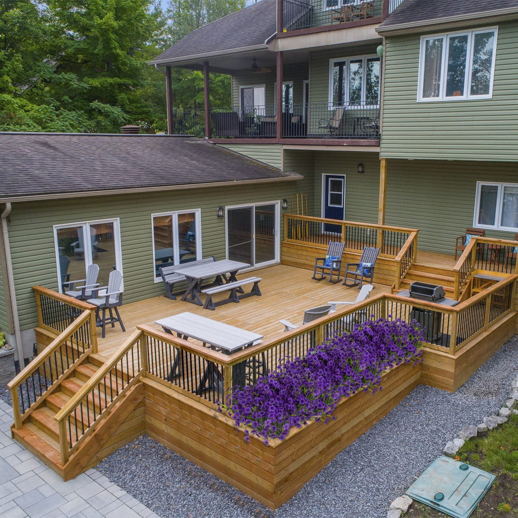 Multi-level wooden deck with outdoor furniture and purple flowers, overlooking a green house.