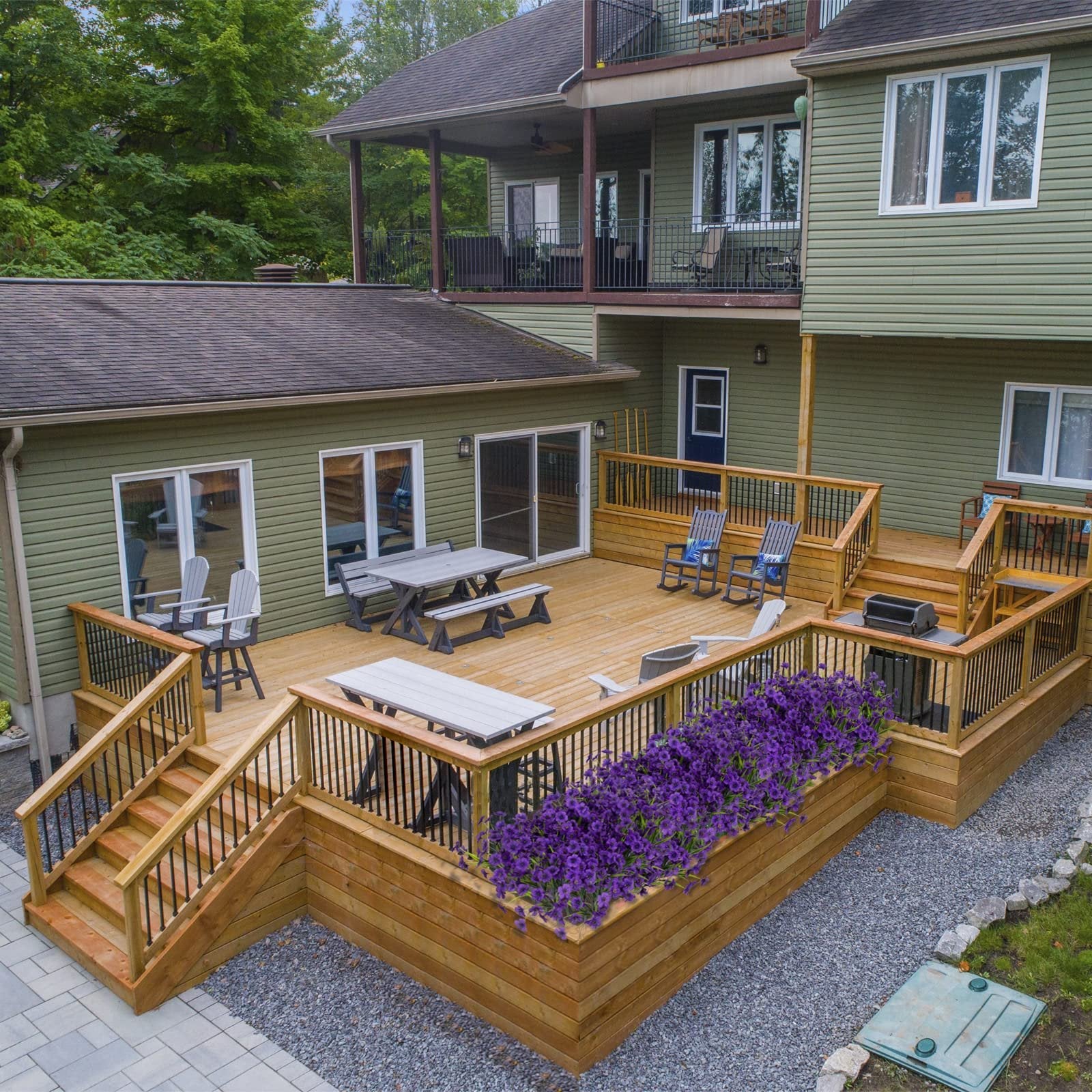 Multi-level wooden deck with outdoor furniture and purple flowers, overlooking a green house.