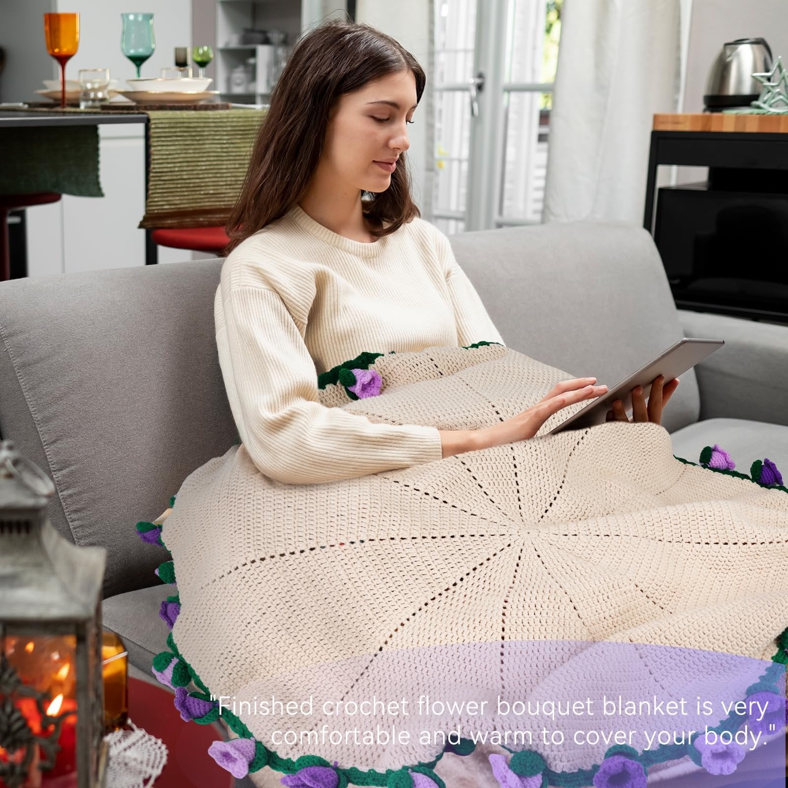 Woman sitting on a couch using a tablet, wrapped in a cream-colored crochet blanket with purple flowers and green leaves.