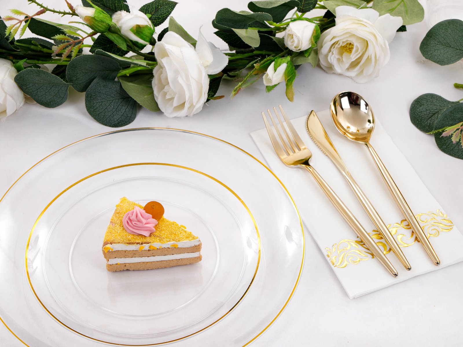 Clear plastic charger plate with gold rim holding a slice of cake, next to gold cutlery on a napkin and floral decor.