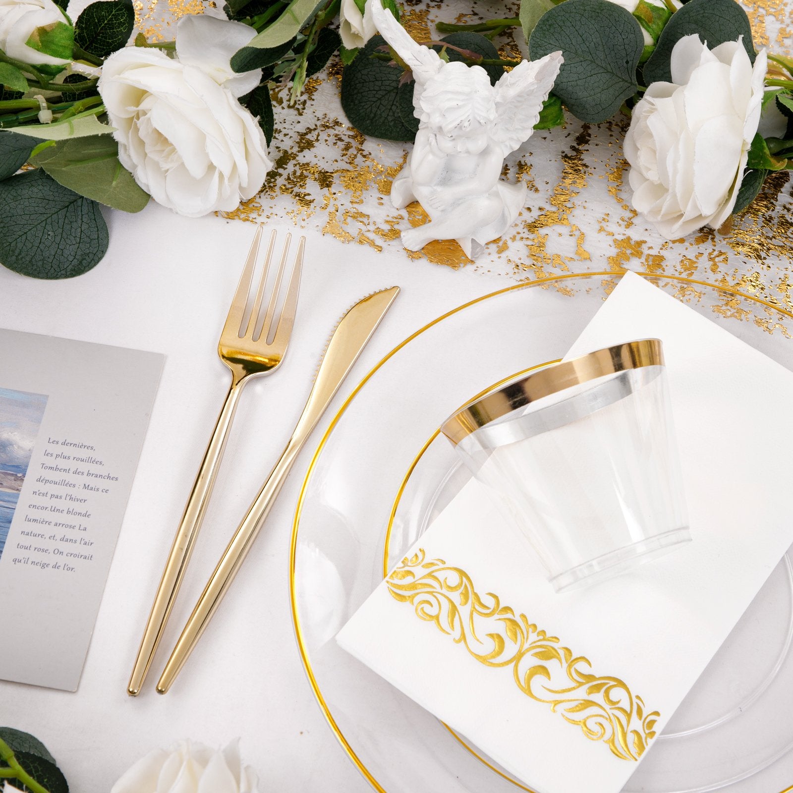 Table setting with gold cutlery, clear plates, and cups, decorated with white roses and gold accents.