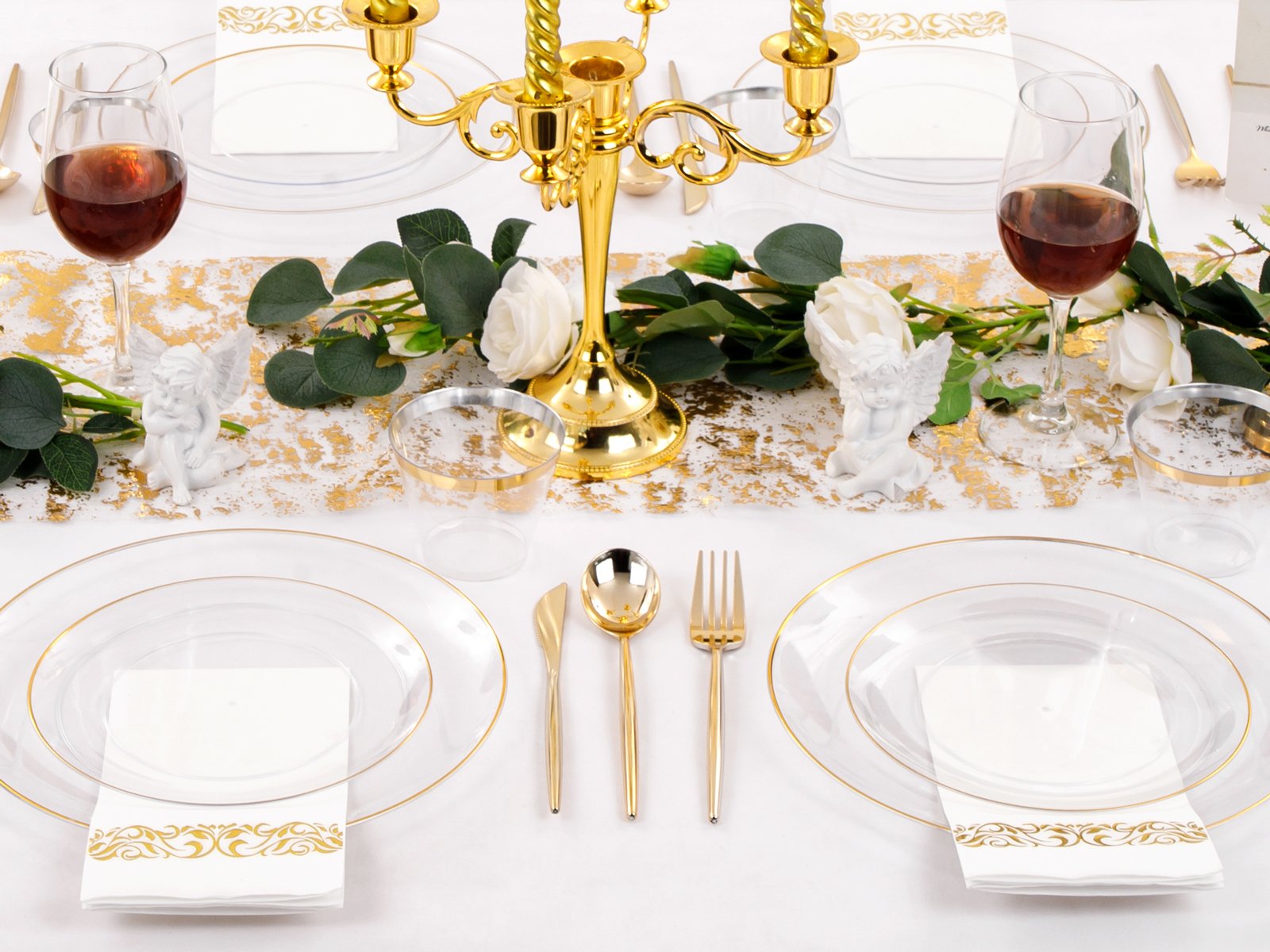 Elegant table setting with clear and gold plastic plates, cutlery, cups, and napkins, adorned with a gold candelabra and white roses.