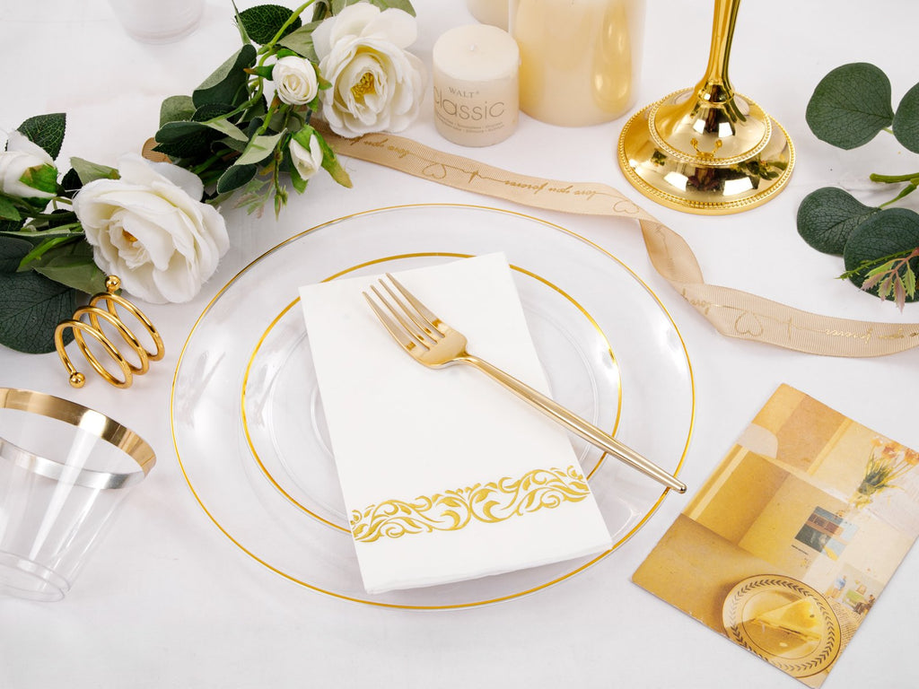 Table setting with clear gold-rimmed plates, white napkins with gold trim, and gold cutlery, surrounded by decorative elements.