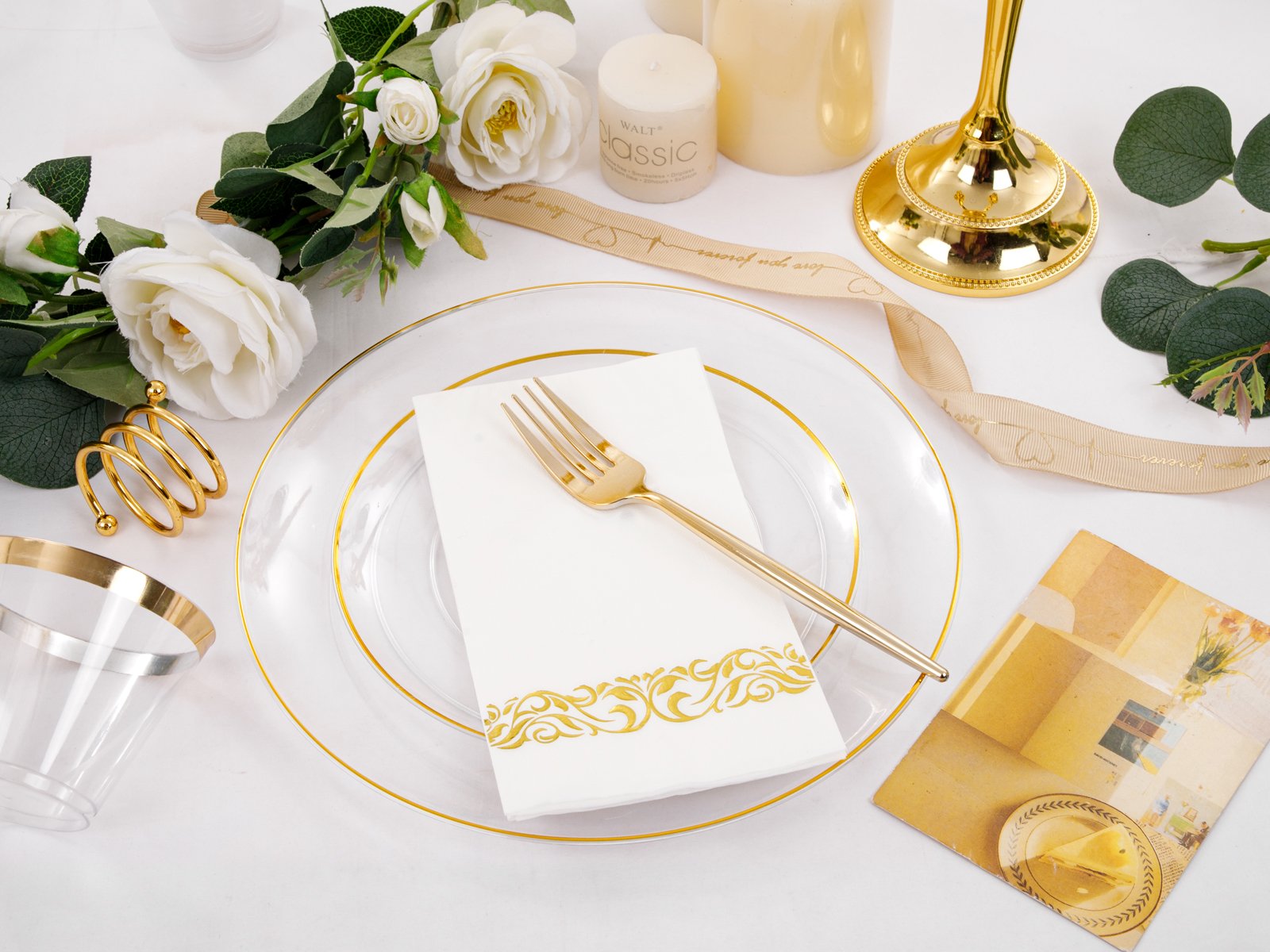 Table setting with clear gold-rimmed plates, white napkins with gold trim, and gold cutlery, surrounded by decorative elements.