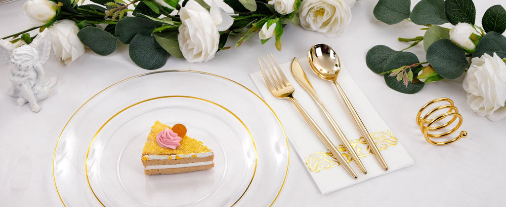 Clear plastic dinner plate with gold rim, set with a slice of cake, gold cutlery on a napkin, and floral decor.