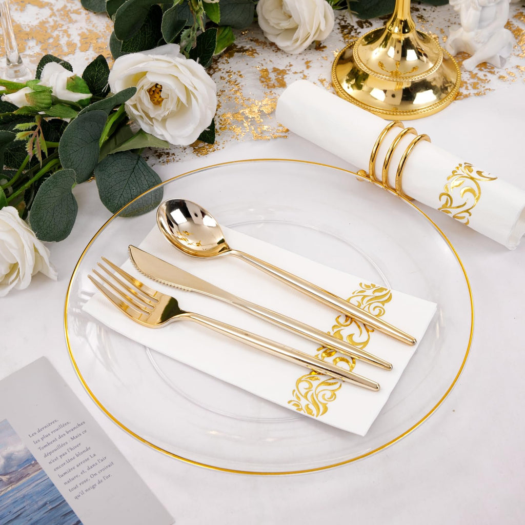 Gold plastic cutlery set on a clear plate with gold trim, next to a white napkin with gold scrollwork and a gold napkin ring.