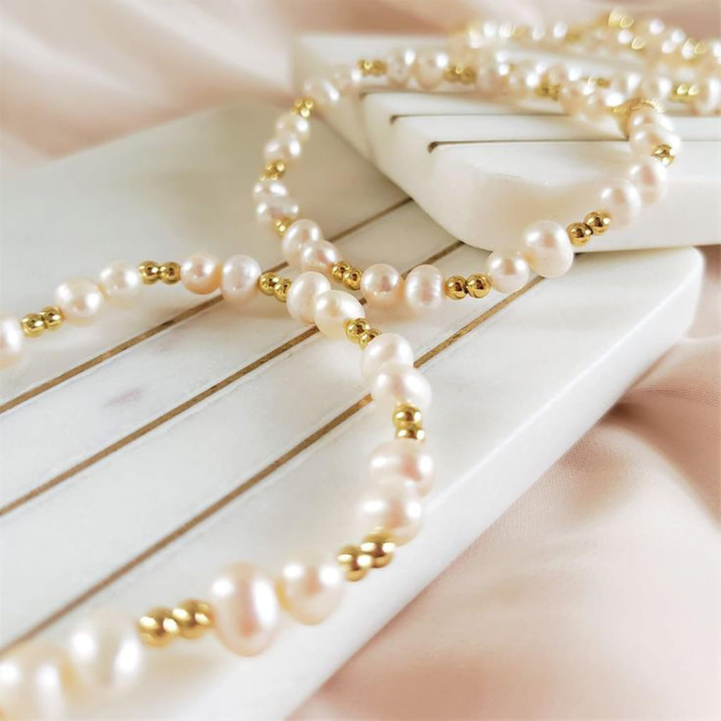 Pearl and gold bead bracelets arranged on a white striped surface with soft pink fabric.