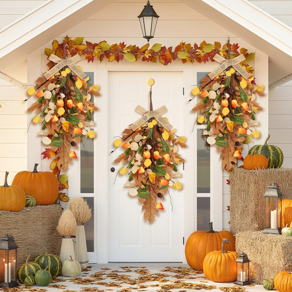 Two fall teardrop swag wreaths with golden eucalyptus and oak leaves hung on a white door, flanked by pumpkins and hay bales.