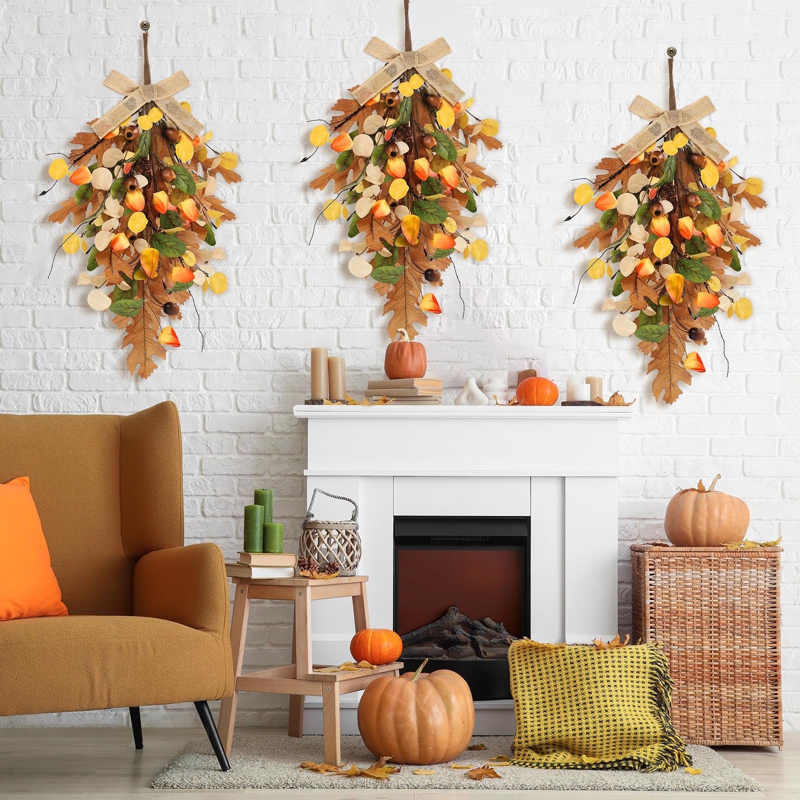 Three fall teardrop swags with golden eucalyptus and oak leaves hanging on a white brick wall above a fireplace.