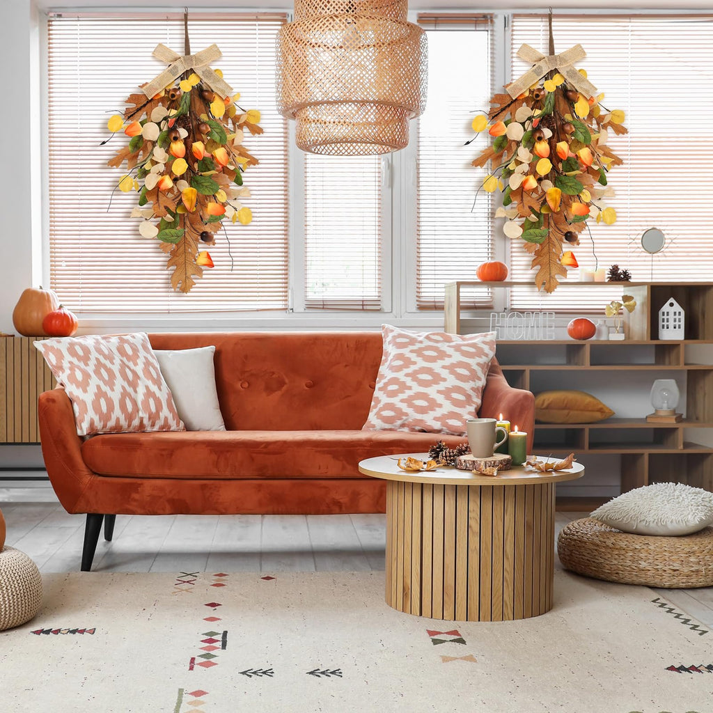 Two fall teardrop swag wreaths with golden eucalyptus and oak leaves hanging on a window with blinds.