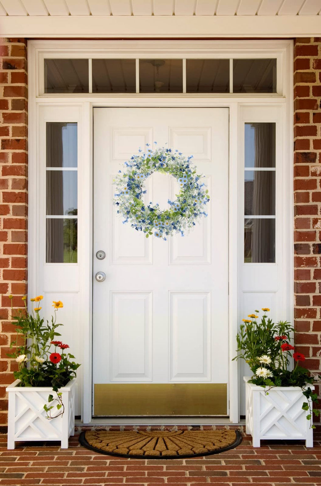 Artificial daisy and wildflower wreath with eucalyptus hanging on a white front door, flanked by planters with flowers.