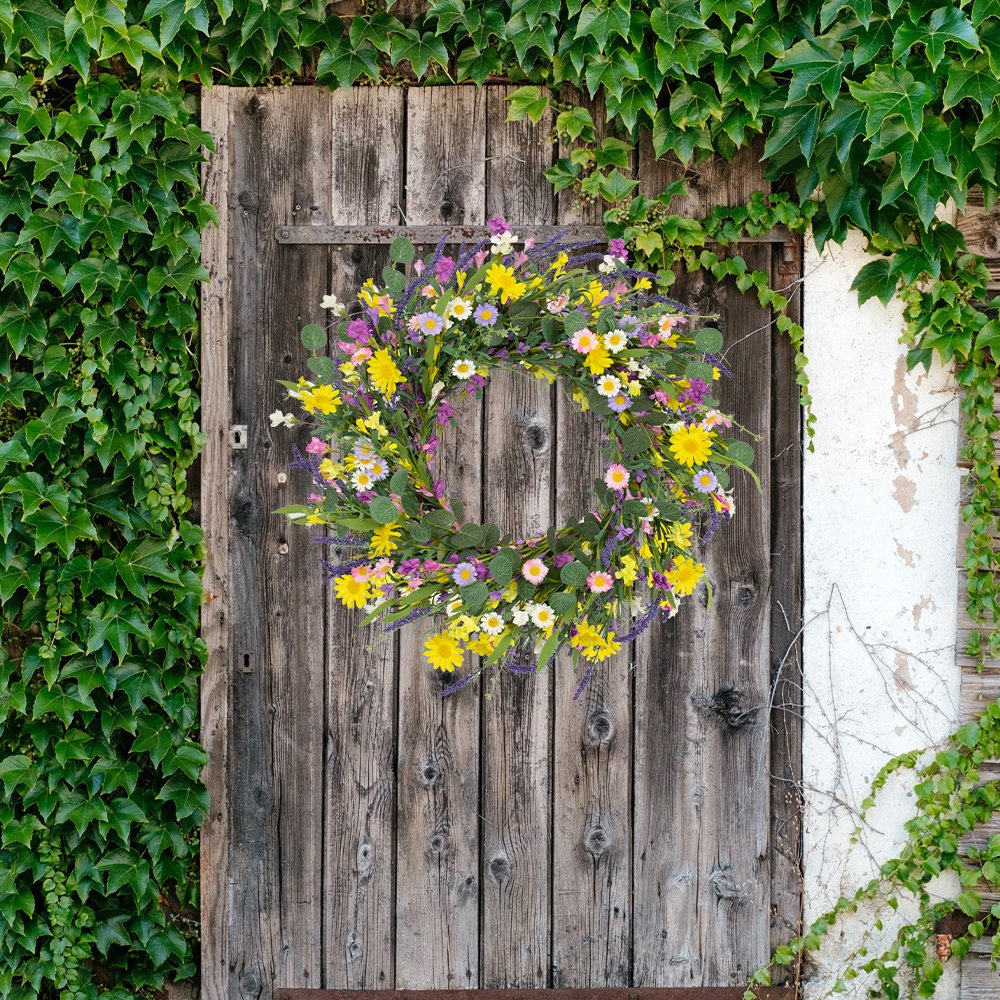 Artificial spring wreath with daisies, lavender, jasmine, and wildflowers on a grapevine base, hanging on a rustic wooden door.