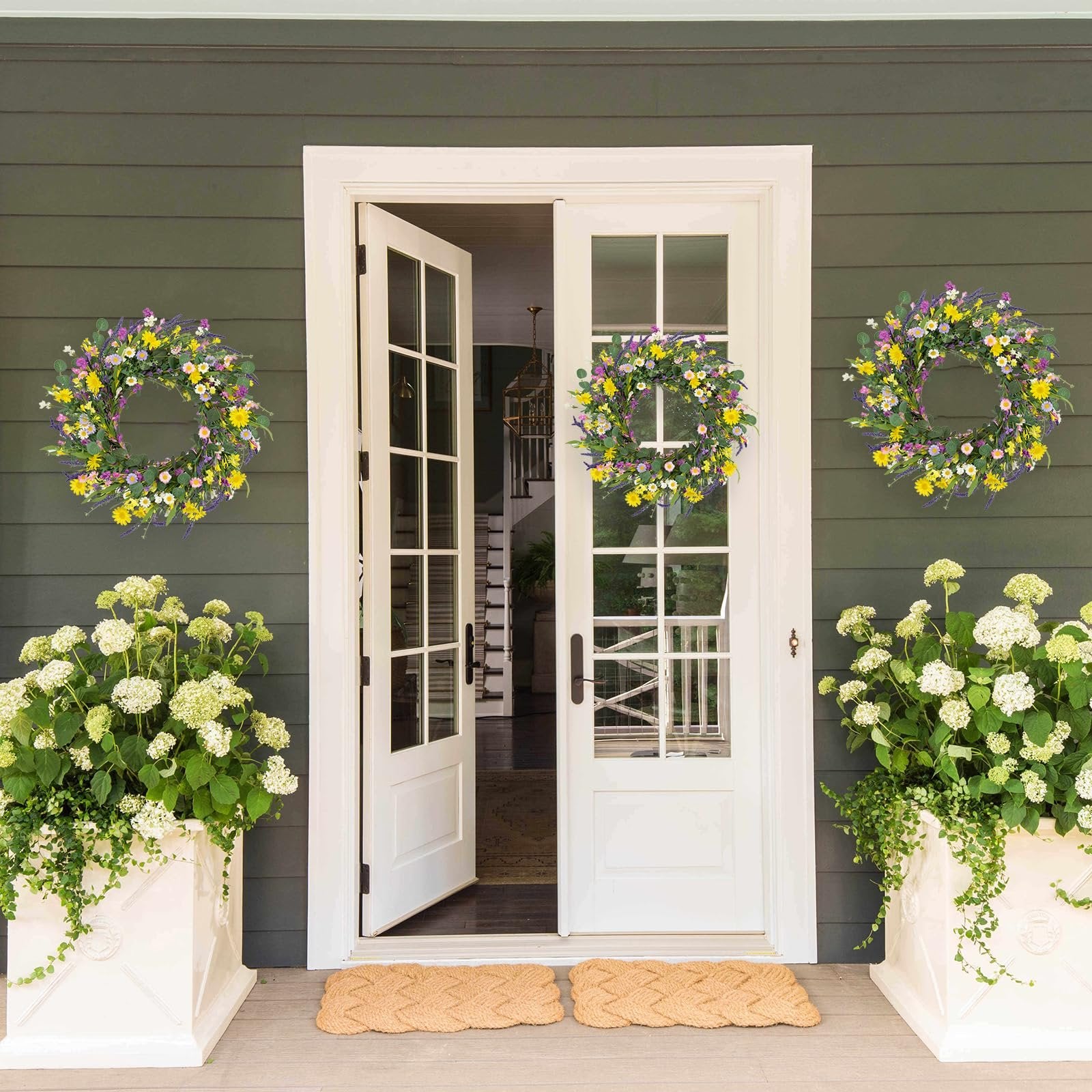 Two artificial spring wreaths with daisies, lavender, and wildflowers hang on a dark green house exterior flanking a white double door.