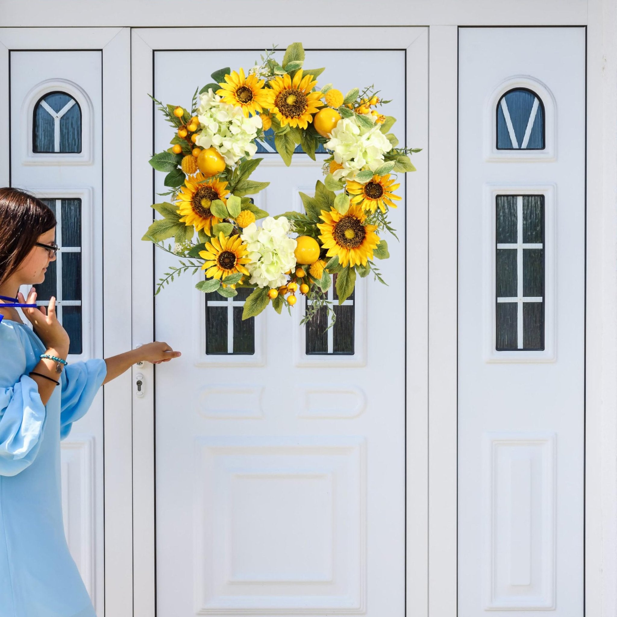 Woman in blue dress reaching for door handle next to a sunflower, hydrangea, lemon, and berry wreath