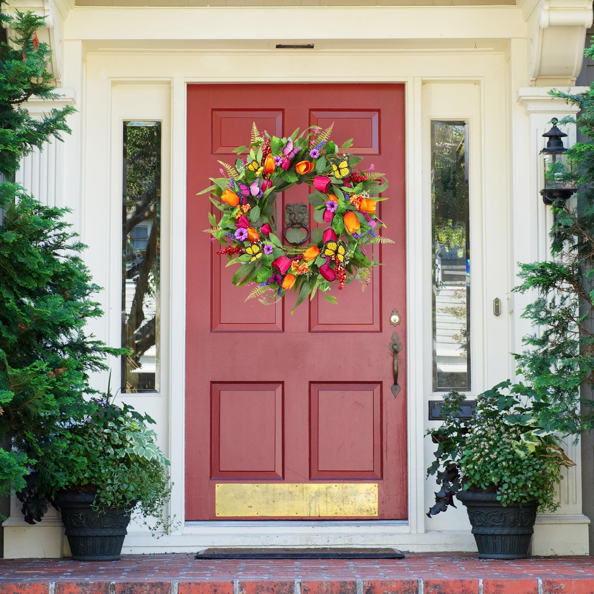 Front door decorated with a colorful tulip and butterfly wreath, flanked by evergreen trees in planters.