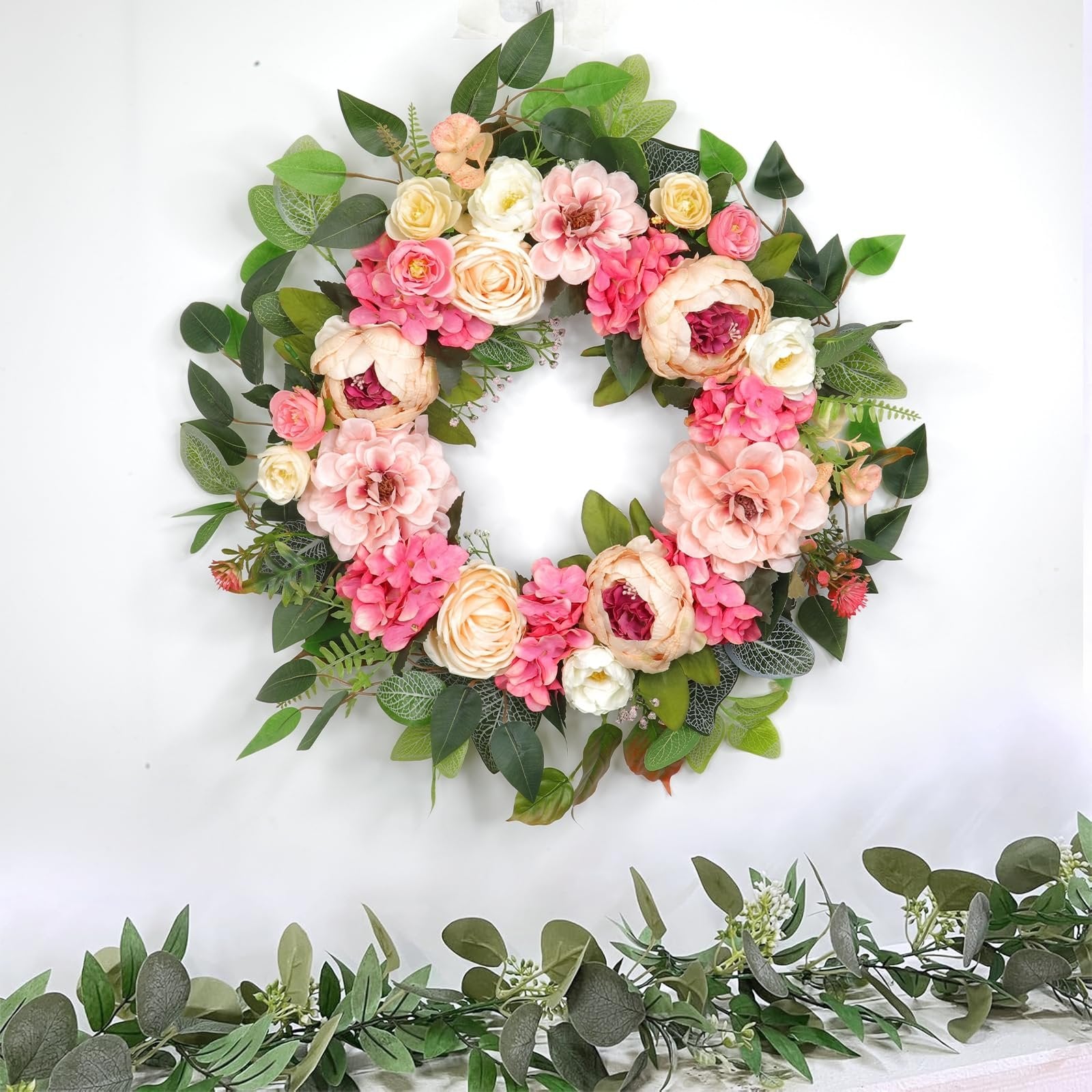 Floral wreath with pink and cream peonies, roses, and hydrangeas, surrounded by green leaves, on a white background with a garland below.