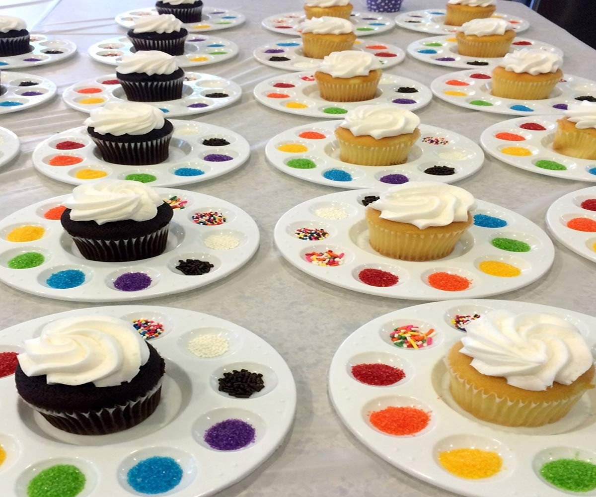 Cupcakes with white frosting on paint palettes filled with colorful sprinkles and sanding sugar.