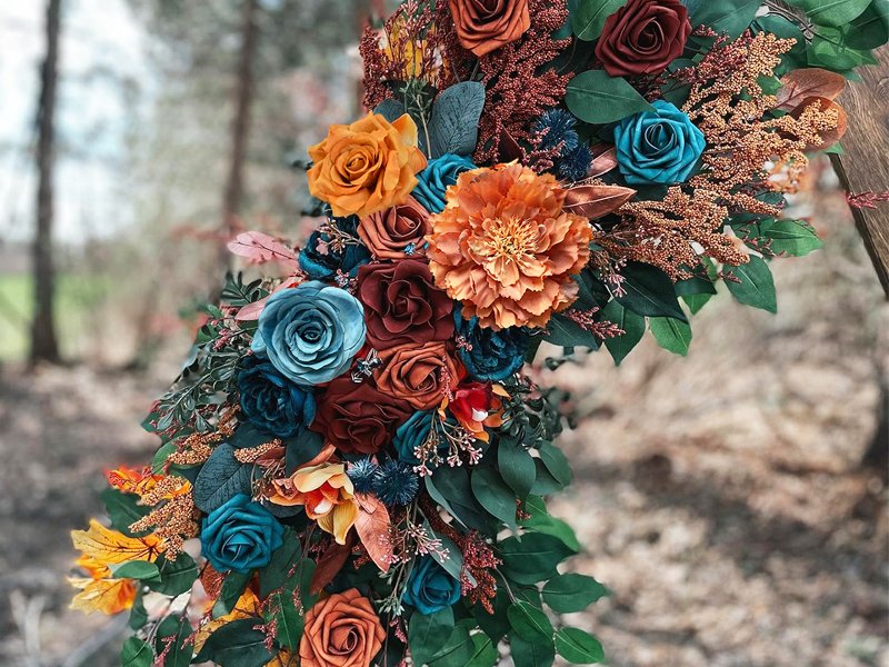 Teal and orange artificial silk flowers arranged on an arch with greenery, set against a blurred outdoor background.
