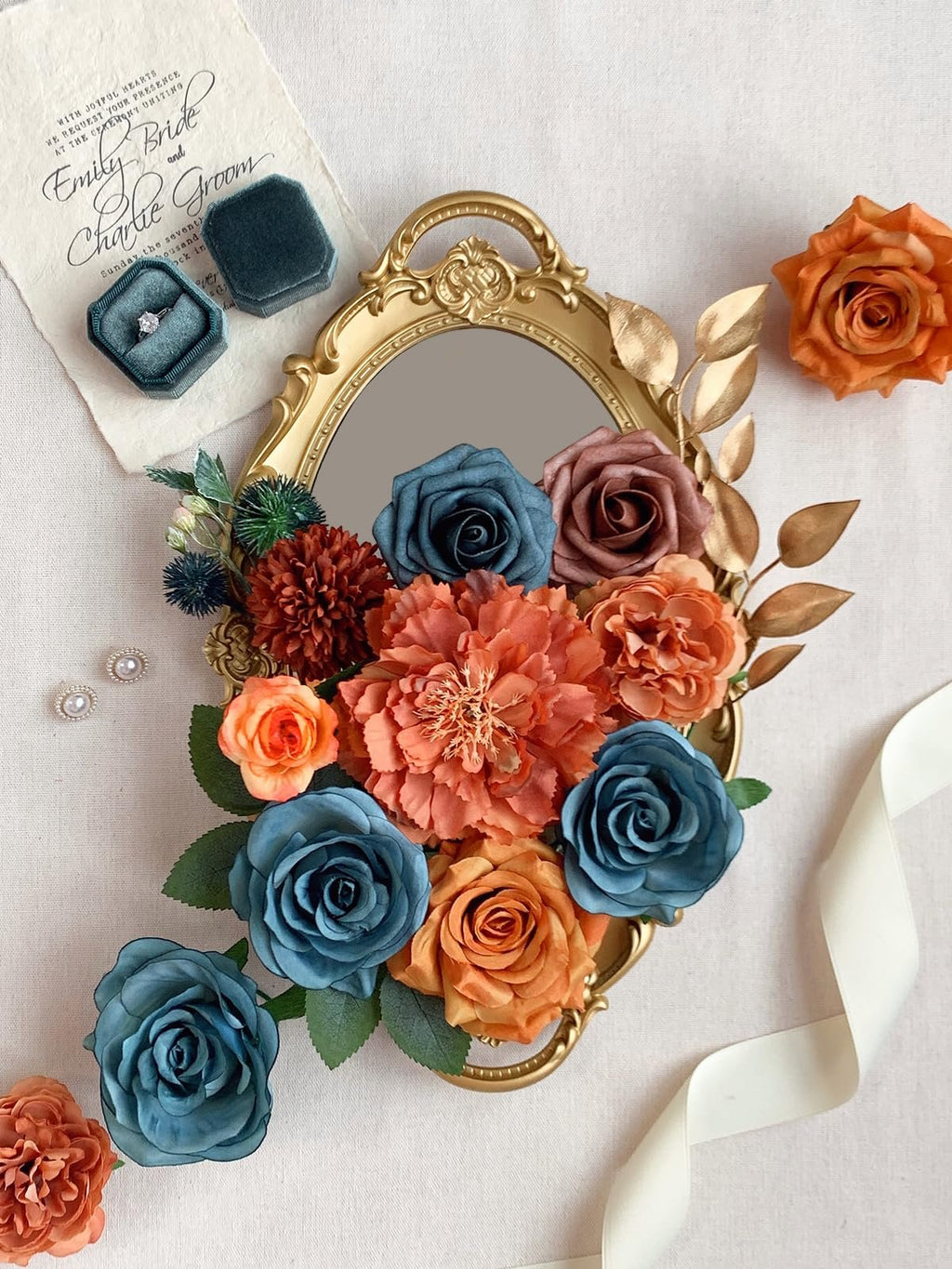 Artificial flowers in teal, orange, and rust hues arranged on a gold ornate tray with a wedding invitation and ring box nearby.