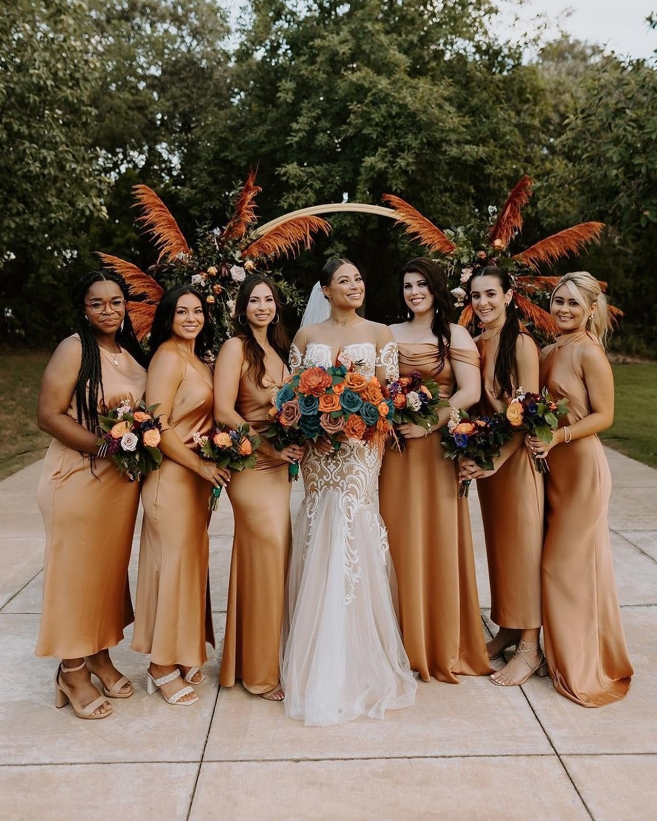 Bride and bridesmaids in gold dresses holding bouquets in front of a wedding arch with pampas grass