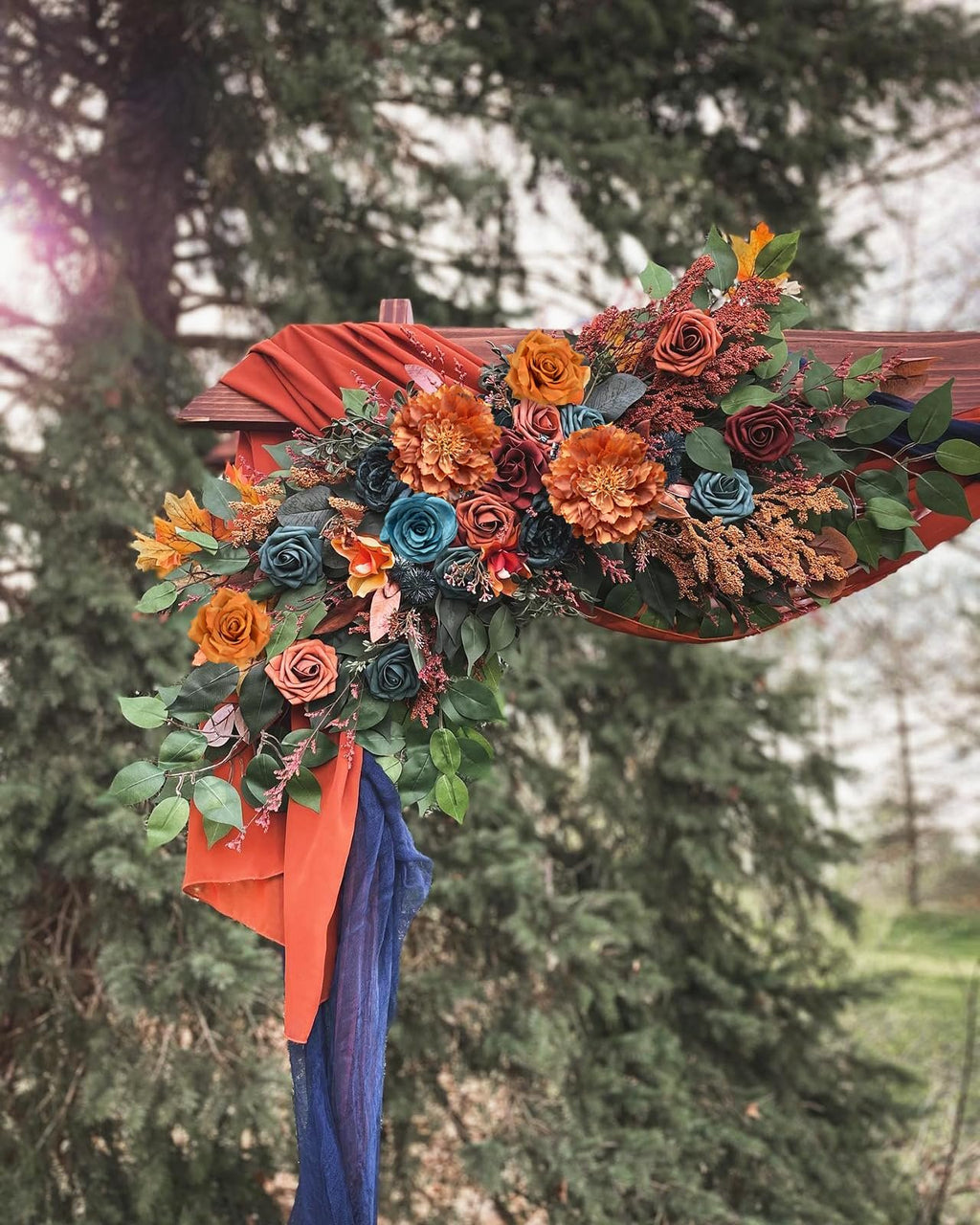 Artificial floral arrangement in teal, orange, and burgundy with greenery draped over a wooden structure outdoors.