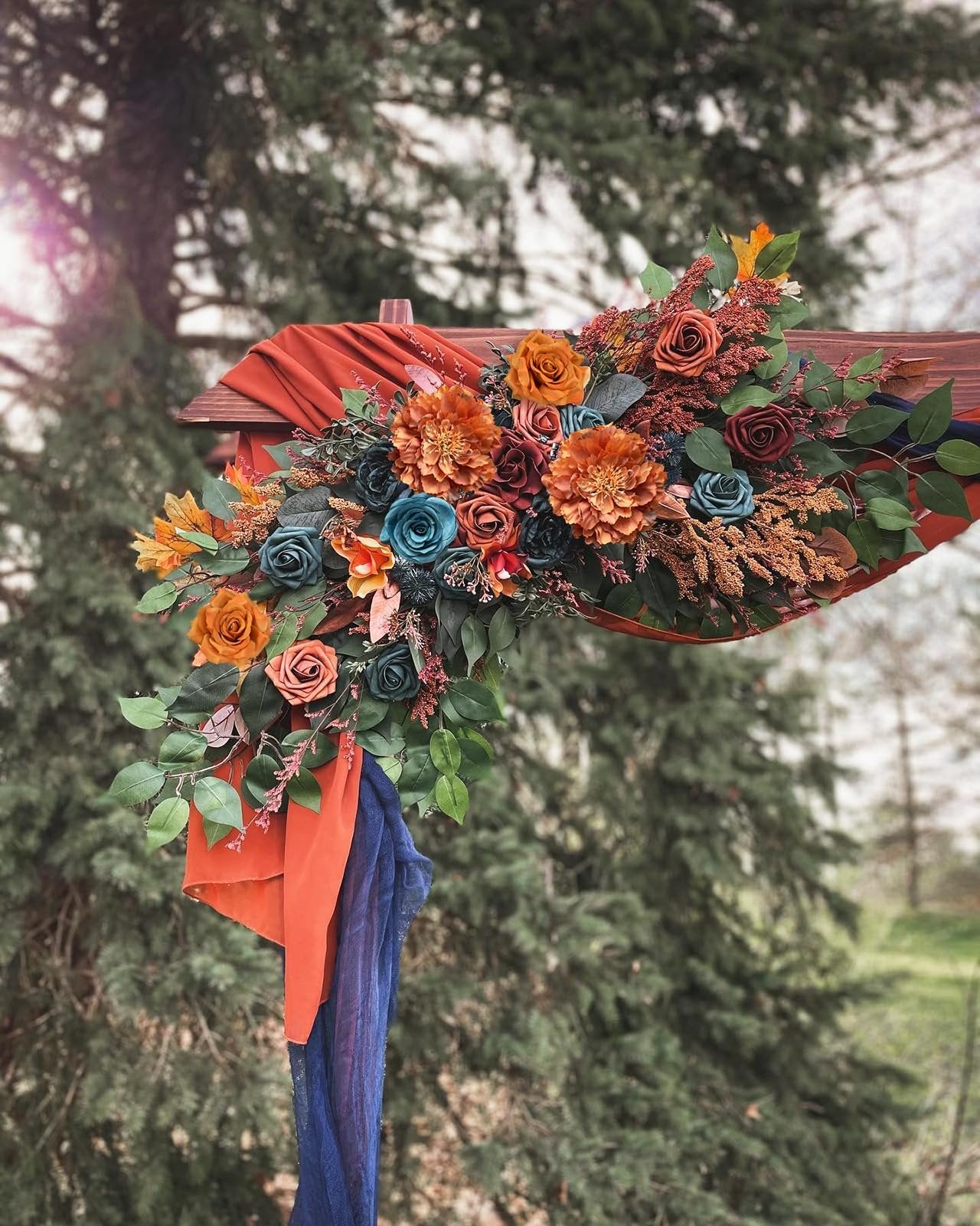 Artificial floral arrangement in teal, orange, and burgundy with greenery draped over a wooden structure outdoors.