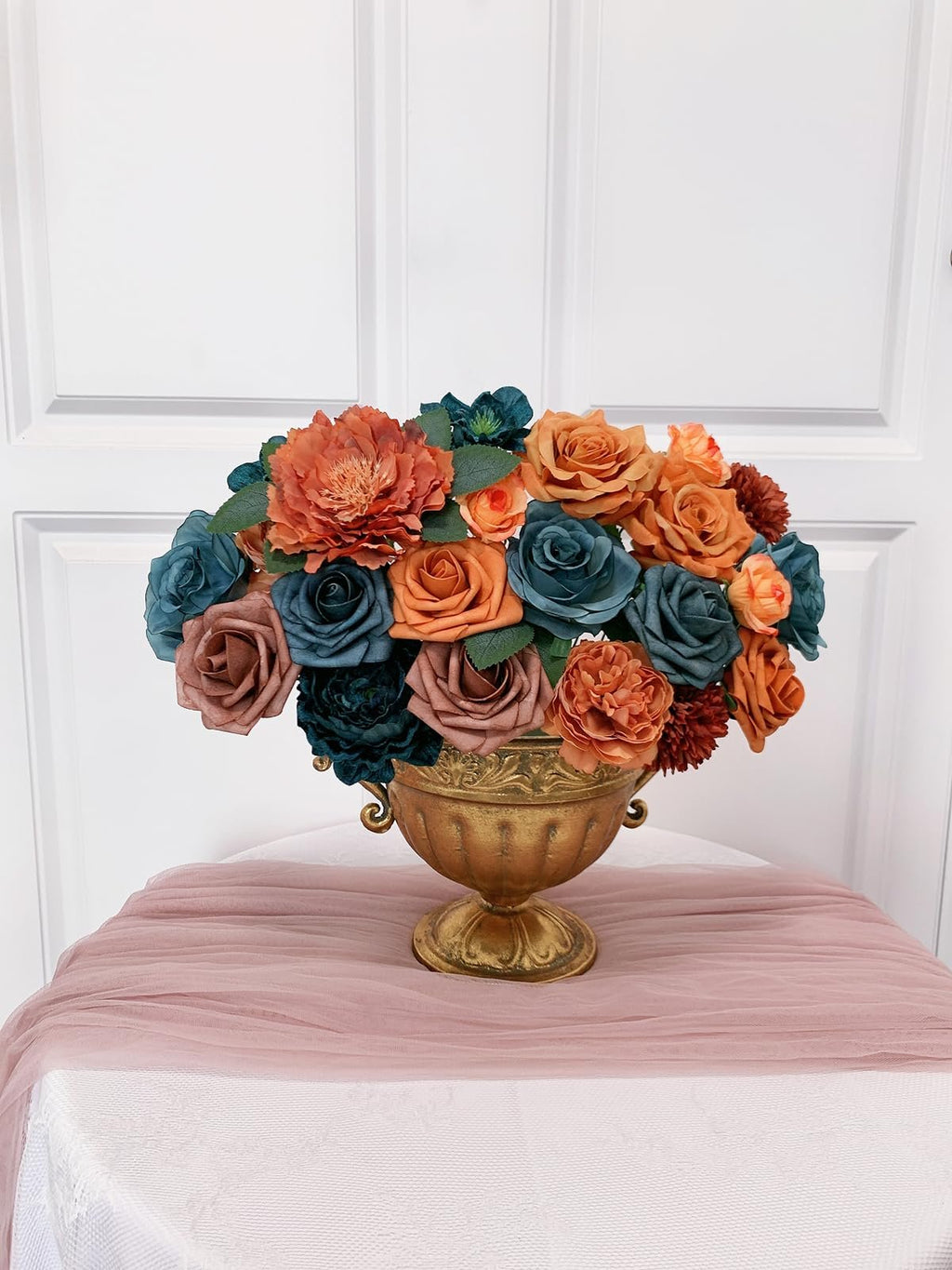 Teal and orange artificial flowers arranged in a gold urn on a table with a pink runner.
