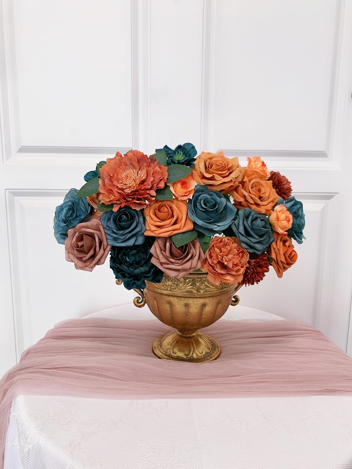 Teal and orange artificial flowers arranged in a gold urn on a table with a pink runner.