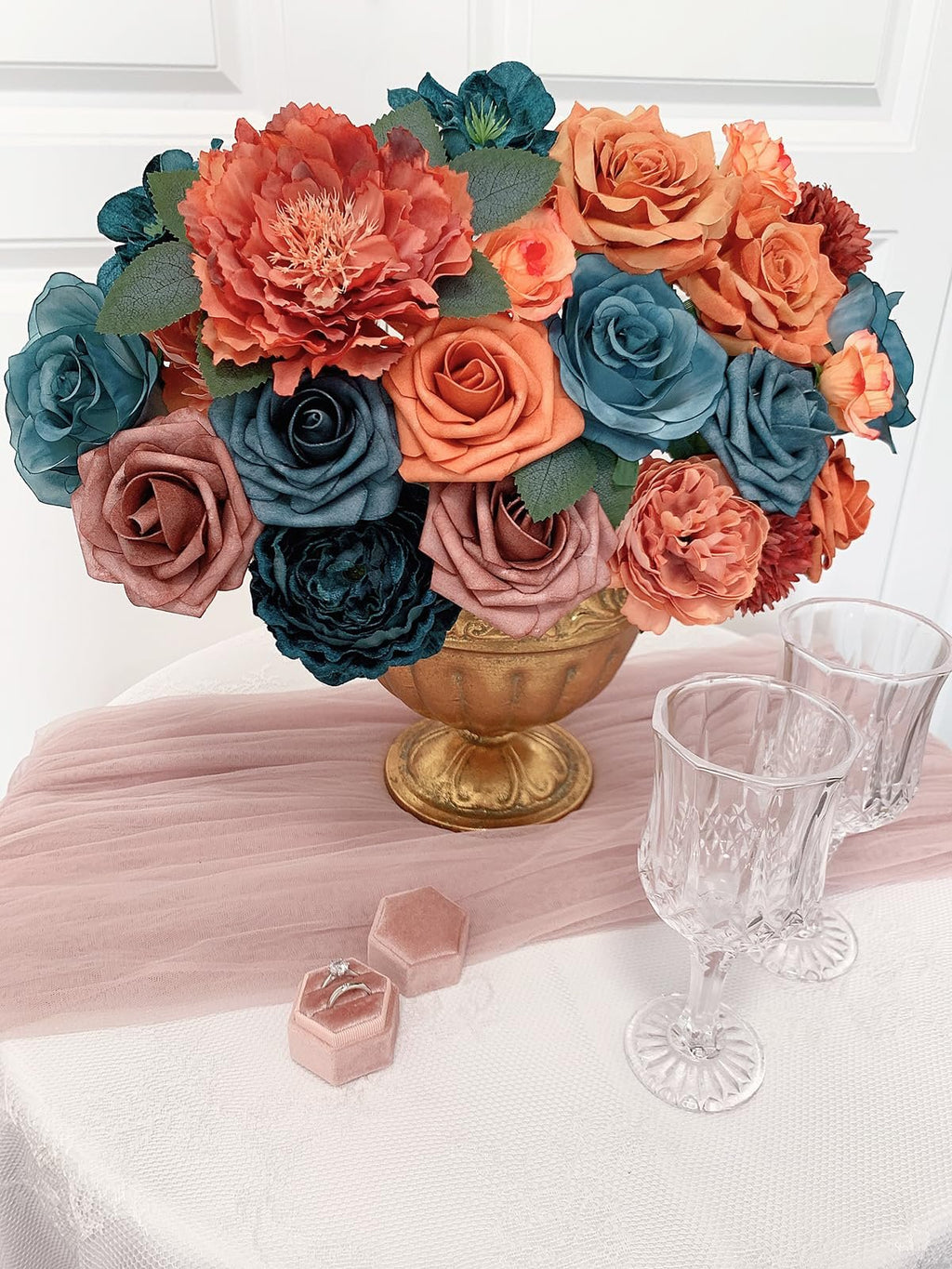 Teal, orange, and rose silk artificial flowers in a gold vase with two crystal glasses and ring boxes on a table.