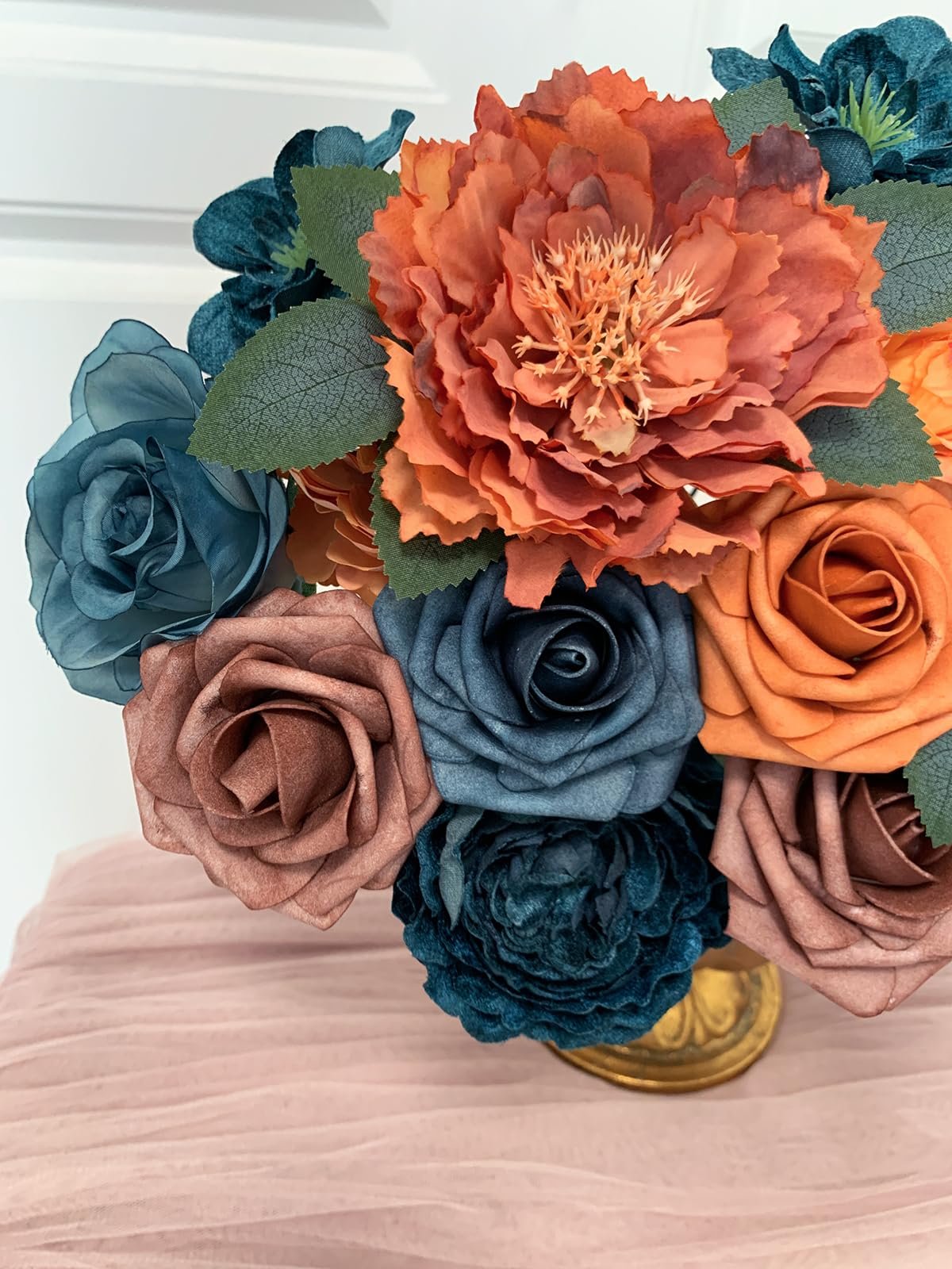 Close-up of a bouquet of artificial silk flowers in teal, orange, and brown hues, arranged in a gold vase on a pink tulle cloth.