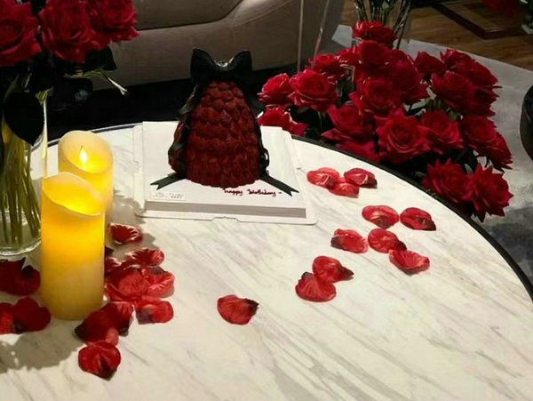 Dark red silk rose petals scattered around a cake and two LED candles, with red roses in the background.