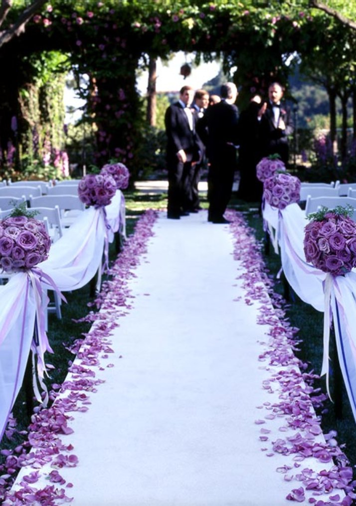 Wedding aisle decorated with purple rose petals and floral arrangements on chairs