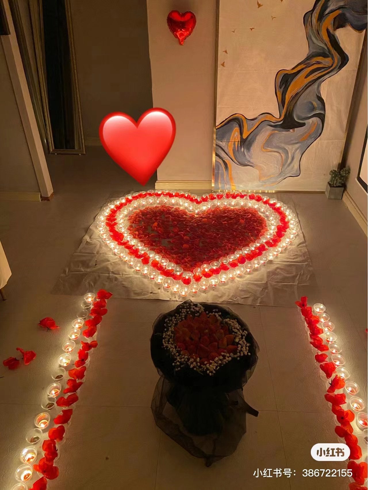 Heart-shaped arrangement of red rose petals and candles, with a bouquet and heart balloons.