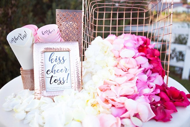 Wedding decor setup with rose petals in ombre shades of pink and white, cones for tossing, and a framed sign.