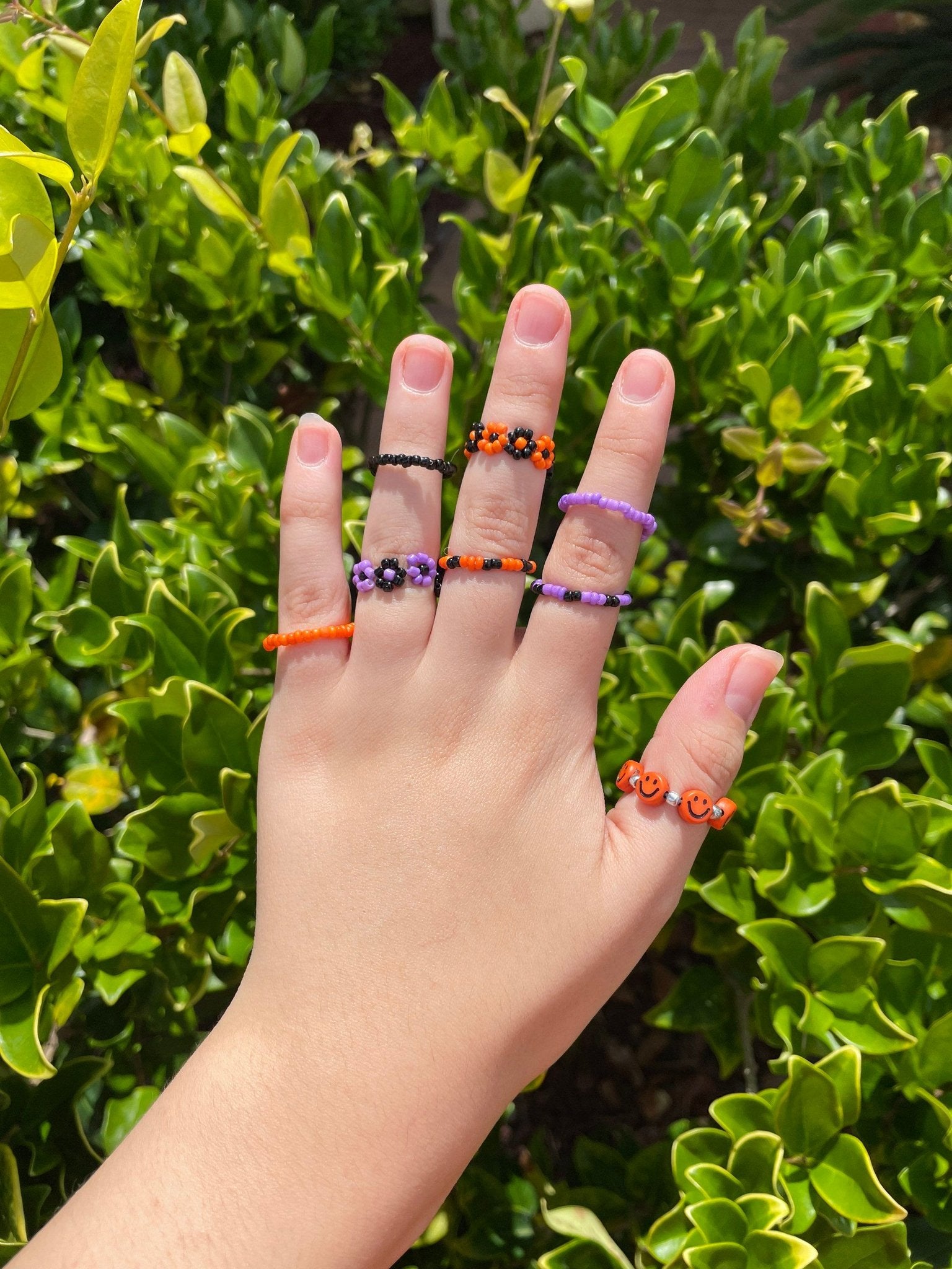 Hand wearing various colorful beaded rings, including orange smiley face rings, purple flower rings, and black and orange rings, against a blurred green leafy background.