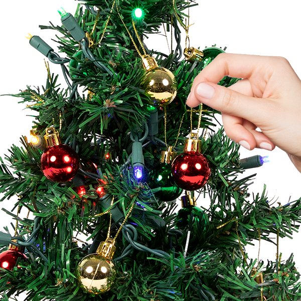 Close-up of a hand decorating a mini Christmas tree with red and gold ornaments and multicolor LED lights.