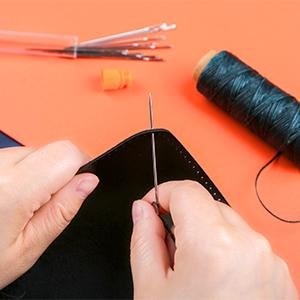 Hands sewing leather with a large eye needle and dark green thread, storage bottle and needles in background