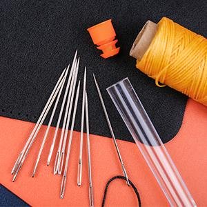 Set of large eye sewing needles with orange thread and a clear storage tube on a black and orange surface.