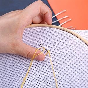 Hand holding a sewing needle and yellow thread through white cross-stitch fabric in an embroidery hoop. Several other needles are visible in the background.