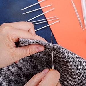 Hands sewing gray fabric with a large eye stainless steel needle, other needles scattered nearby.