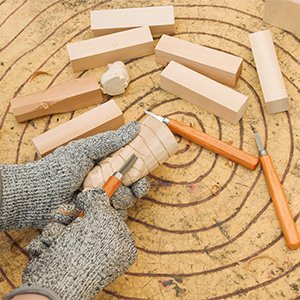 Hands wearing protective gloves carve a wooden cylinder with wood carving tools on a circular wooden surface.